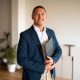 A mid-adult Hispanic male real estate agent smiles engagingly, clad in professional attire with a folder in hand, standing inside a modern office environment.