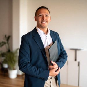 A mid-adult Hispanic male real estate agent smiles engagingly, clad in professional attire with a folder in hand, standing inside a modern office environment.